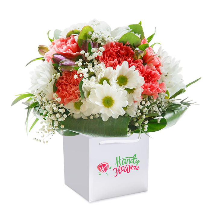 An extravagantly arranged bouquet, poised with pride in a flawless square white gift bag, brings a breath of Wennington's charm to life through flowers. The composition showcases a lush gathering of soft coral-pink and lively peach carnations-their intricate, ruffled petals layered for maximum fullness and texture-balanced by a selection of brilliant white daisies, each with a striking lime-green center radiating vitality. Wispy sprigs of white baby's breath, like delicate clouds, tumble between the showier blooms, softening the arrangement and adding an air of innocent celebration. Framing the ensemble is abundant, verdant foliage, including wide, lustrous green leaves which create a natural cradle at the bouquet's base, supporting the rounded, symmetrical form and making the whole ensemble appear both substantial and inviting. Nestled discreetly along one edge, partially opened buds with hints of purple and blush quietly allude to new beginnings and hidden surprises. Designed for effortless delivery-whether to a neighbour near Wennington Green or as an elegant table accent for a family celebration-the pristine white bag features strong ribbon handles and a tasteful local branding mark. The bouquet is illuminated with gentle, diffused light, rendering each blossom's nuance, lushness, and freshness with clarity. The composition feels tailor-made for gift-giving occasions-birthdays, anniversaries, or spontaneous tokens of affection-served with the personal touch only a Wennington florist can provide, blending local artistry with timeless floral beauty.